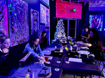 Group painting workshop under purple-blue blacklight in an indoor art studio — long table with canvases and art supplies, participants creating neon artwork, vibrant abstract paintings on the walls and a lit Christmas tree in the background.