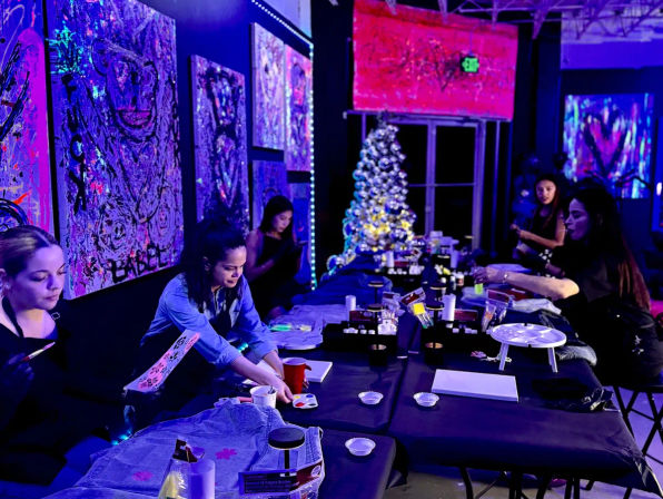 Group painting workshop under purple-blue blacklight in an indoor art studio — long table with canvases and art supplies, participants creating neon artwork, vibrant abstract paintings on the walls and a lit Christmas tree in the background.