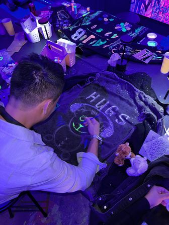 Overhead view of a person painting a neon bear and the word "HUGS" on a denim jacket under UV lights at an indoor glow-in-the-dark art workshop, surrounded by paint supplies, stencils, and plush toys.