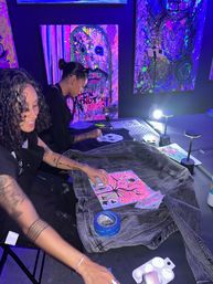 Two people painting a denim jacket under blacklight in a neon-lit art studio, using fluorescent stencils, tape and bright abstract canvases