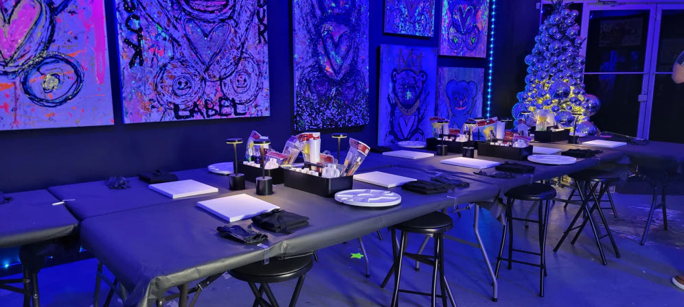 UV-lit art studio set for paint night with long tables, blank canvases, palettes and paint kits, neon purple-blue lighting, colorful abstract murals and a silver ornament tree.
