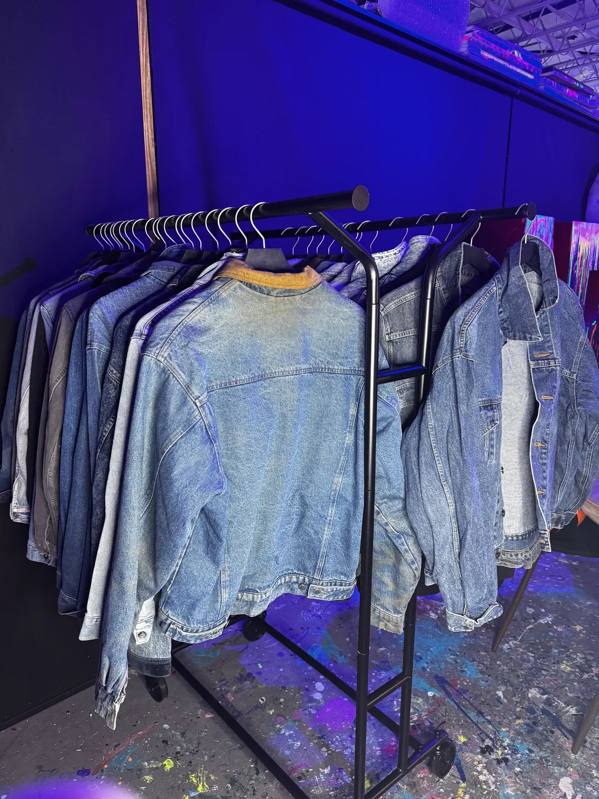 Row of vintage-style denim jackets hanging on a black clothing rack under purple neon lights in a paint-splattered art studio