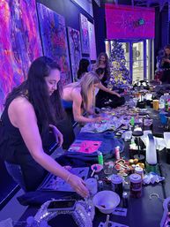 Neon-lit art studio paint party: people gathered around a long table decorating denim jackets and canvases with bright paints, glitter and stickers, colorful abstract wall art and a silver Christmas tree by a storefront window.
