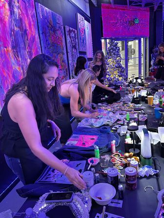 Neon-lit art studio paint party: people gathered around a long table decorating denim jackets and canvases with bright paints, glitter and stickers, colorful abstract wall art and a silver Christmas tree by a storefront window.