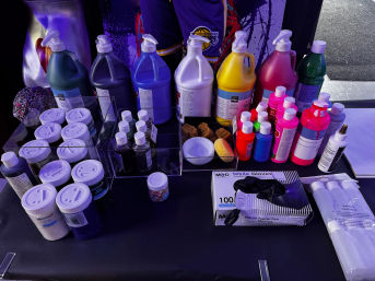 Colorful craft table with gallon and squeeze bottles of acrylic paint, small glitter jars, sponges, mixing bowls and a box of nitrile gloves on a black work surface under purple lighting