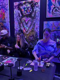 Three people painting at a neon-lit art studio paint night, working with palettes, brushes and a water bottle in front of a large colorful abstract bear canvas featuring a heart motif.
