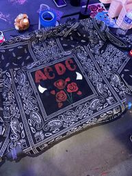 Black paisley bandana-style cloth on a craft table with a central red-lettered graphic and three painted red roses, surrounded by blue painter’s tape, glitter, a small plush toy and a water bottle.