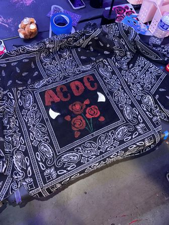 Black paisley bandana-style cloth on a craft table with a central red-lettered graphic and three painted red roses, surrounded by blue painter’s tape, glitter, a small plush toy and a water bottle.