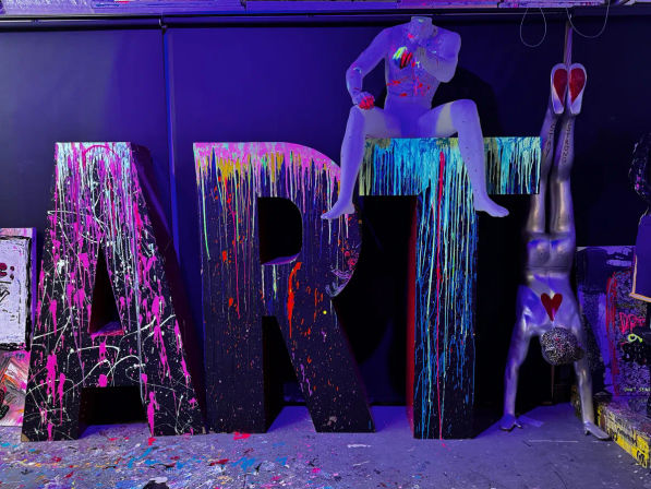 Contemporary art installation in a dark gallery: giant letters spelling ART covered in neon paint splatter, a painted mannequin perched on the T, and a silver upside-down mannequin with a red heart doing a handstand under purple/blacklight with paint-splattered floor.