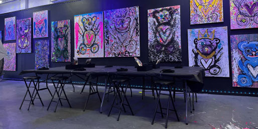 Neon-lit warehouse art studio featuring playful abstract teddy-bear canvases on a black wall, with a long black-covered table and folding stools set up for a group painting session.