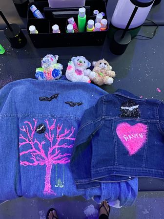 DIY craft table with two custom-painted denim jackets: a larger jacket decorated with a neon pink glitter tree and black bird silhouettes, and a smaller jacket featuring a bright pink glitter heart with white stenciled text; three colorful painted plush teddy bears, bottles of neon paint and glitter scattered around, sandals peeking at the bottom.