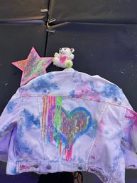 Kids' tie-dye denim jacket with glittery rainbow heart and neon paint drips, laid on a black mat next to a sparkly pink star craft and a small plush bear — colorful DIY art project