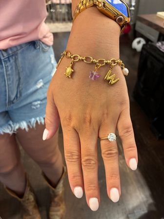 Close-up of a hand with a sparkling solitaire engagement ring, white manicure and gold charm bracelet with a turtle, purple butterfly, letter W and pearl; gold watch, denim cutoff shorts and cowboy boots suggest a casual western summer look.