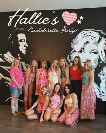 Group of ten women smiling at an indoor bachelorette party, posing in front of a large black wall mural with stylized portrait art and script signage; many wearing pink, red, gingham and denim outfits and cowboy boots.