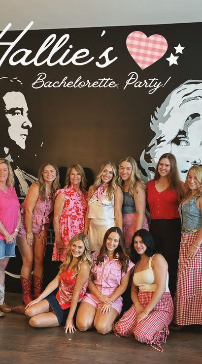 Group of ten women smiling at an indoor bachelorette party, posing in front of a large black wall mural with stylized portrait art and script signage; many wearing pink, red, gingham and denim outfits and cowboy boots.
