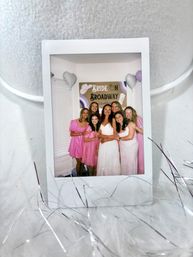 Polaroid-style photo of a smiling bride in a white dress surrounded by bridesmaids in pink dresses at an indoor bachelorette party with a 'Bride on Broadway' sign and heart-shaped balloons
