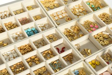 Close-up of a beige velvet jewelry organizer tray filled with assorted gold-tone charms and pendants—stars, hearts, suns, crosses, enamel and pearl accents—neatly sorted in compartmented retail display.