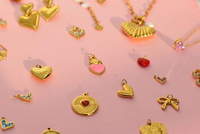 Scattered gold-tone heart and bow jewelry charms with pink and red enamel accents on a pastel pink background