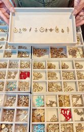 Close-up of hands holding a jewelry display tray filled with assorted gold charms and pendants neatly organized in lined compartment trays, with red and turquoise enamel accents.