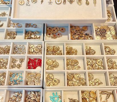 Close-up of hands holding a jewelry display tray filled with assorted gold charms and pendants neatly organized in lined compartment trays, with red and turquoise enamel accents.
