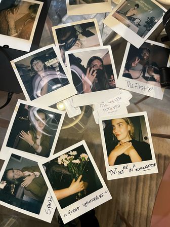 Polaroid instant photos scattered on a glass tabletop — candid portraits of friends, handwritten captions, and a small bouquet.