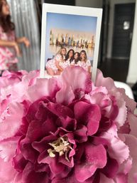 Pink and magenta fabric peony centerpiece dusted with glitter holding an instant photo of four friends in matching pink pajamas posed on a bed with a city skyline painting — indoor party table display.
