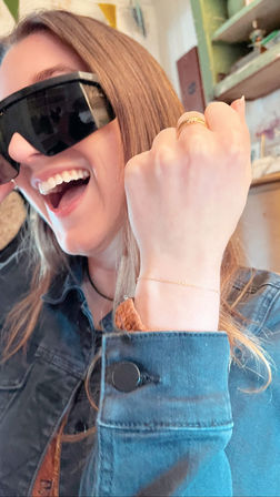 Smiling woman in oversized black sunglasses and denim jacket, laughing with fist raised to show gold rings and a delicate chain bracelet in a cozy cafe-style interior.