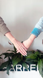 Two hands touching above a leafy indoor plant against a white wall, sporting a sparkling ring, dainty bracelets and pale manicured nails — cozy lifestyle moment.