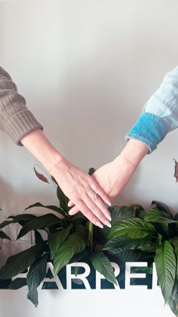 Two hands touching above a leafy indoor plant against a white wall, sporting a sparkling ring, dainty bracelets and pale manicured nails — cozy lifestyle moment.