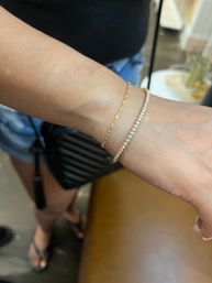 Close-up of a wrist wearing a delicate gold paperclip chain and a sparkling diamond tennis bracelet, jewelry close-up with casual summer outfit and crossbody bag blurred in the background.
