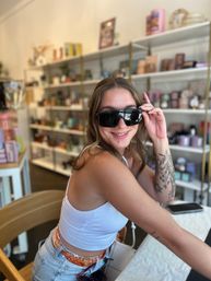 Smiling woman trying on black sunglasses inside a bright boutique gift shop, wearing a white tank top and denim, showing a floral arm tattoo with shelves of candles and home decor blurred behind her.