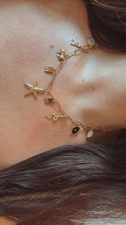 Close-up of a gold charm paperclip-chain necklace with starfish, snake, flower and black enamel eye charms resting on a collarbone with brown hair