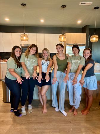 Six smiling women in casual loungewear posing in a modern open-plan kitchen with pendant lights and wood floors, holding their hands forward to showcase rings and bracelets.