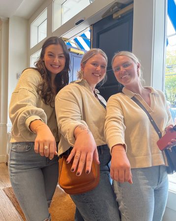 Three smiling women in light sweaters and jeans at a sunlit storefront with a blue-striped awning, extending hands to show rings and dark nail polish — casual downtown shopping moment.