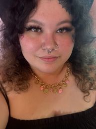Close-up portrait of a smiling person with curly dark hair, septum and nostril rings, dramatic lashes, small heart cheek and spiderweb forehead tattoos, wearing a chunky gold charm necklace with pink hearts and a black top.
