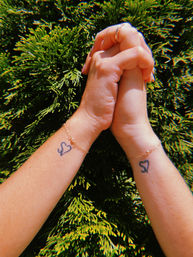 Sunlit close-up of two clasped hands with matching heart wrist tattoos and delicate gold chain bracelets against a lush green hedge