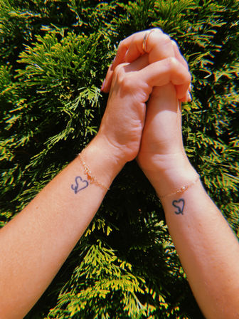 Sunlit close-up of two clasped hands with matching heart wrist tattoos and delicate gold chain bracelets against a lush green hedge