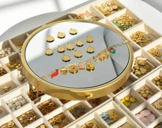 Round mirror tray showcasing a gold charm bracelet and alphabet coin pendants atop a compartmental jewelry organizer filled with assorted gold and colorful enamel charms