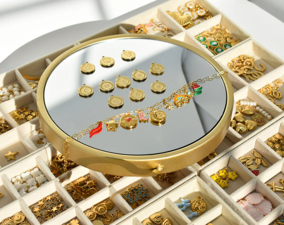 Round mirror tray showcasing a gold charm bracelet and alphabet coin pendants atop a compartmental jewelry organizer filled with assorted gold and colorful enamel charms
