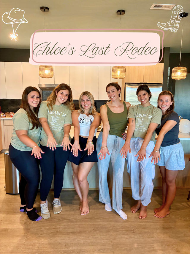 Six friends in a modern kitchen under a pink "Chloe's Last Rodeo" sign, smiling and showing engagement rings — casual bachelorette party scene with pendant lights and wood floors