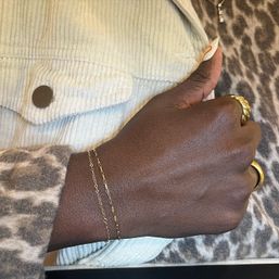 Close-up of a hand wearing delicate gold chain bracelets and chunky gold rings with a long almond-shaped nail, resting on leopard-print fabric and a cream corduroy jacket.