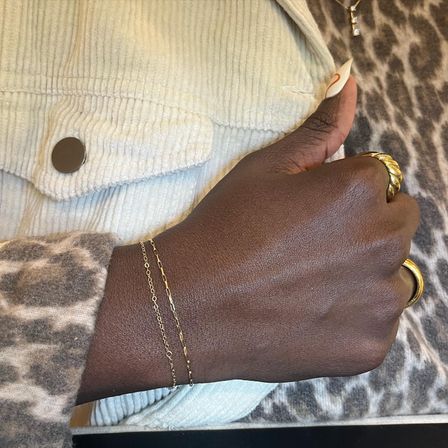 Close-up of a hand wearing delicate gold chain bracelets and chunky gold rings with a long almond-shaped nail, resting on leopard-print fabric and a cream corduroy jacket.