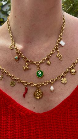 Close-up of layered gold charm necklaces — sun, snake, four-leaf clover, pearl, green flower and red horn charms — worn on a neck above a red knit sweater