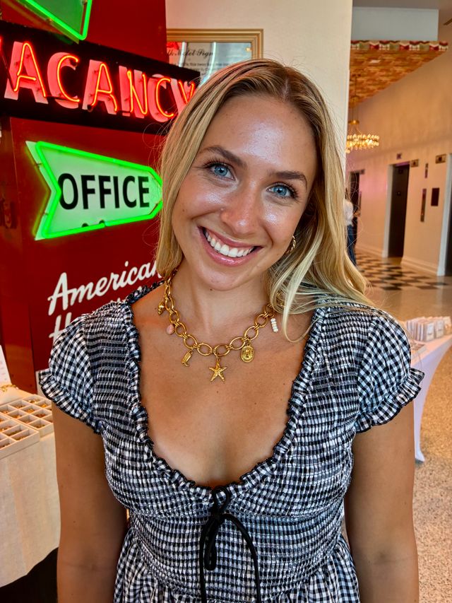 Smiling woman in a black-and-white gingham dress and gold charm necklace posing in a retro hotel lobby with neon "VACANCY" and green "OFFICE" signs and a checkerboard floor.
