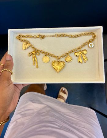 Gold charm necklace with hearts, bows, shell and cross charms on a cream velvet display tray held by a hand — boutique jewelry product photo