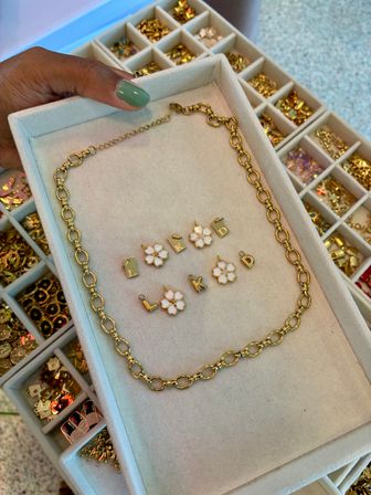 Close-up of a gold link necklace and white enamel flower charms with letter initial pendants arranged on a soft jewelry display tray, held by a hand with green nail polish; compartmented case of gold charms visible in the background.