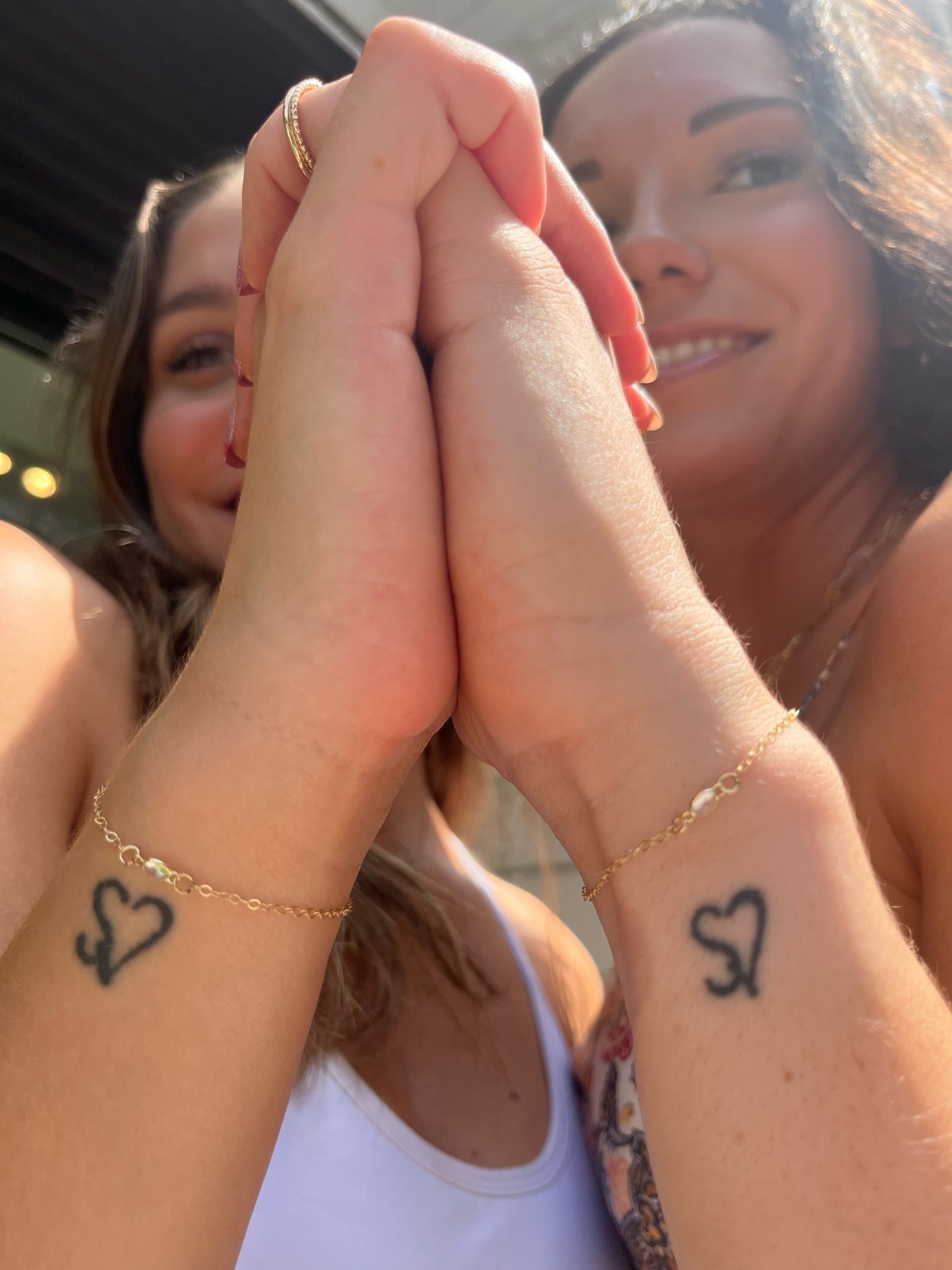 Sunlit close-up of two friends pressing palms together, showing matching heart wrist tattoos and delicate gold chain bracelets while smiling outdoors.
