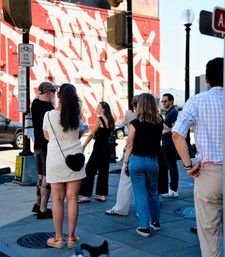 Group of pedestrians waiting at a sunny downtown crosswalk in front of a bold red-and-white mural, with bike-use signs, traffic light and lamppost visible on the city sidewalk.