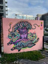 Vibrant outdoor street art mural on a pink building wall: a purple-skinned woman with space-bun hair lounging amid two neon-striped tigers in teal, pink and yellow, with city buildings in the background.