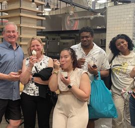 Five adults smiling and sampling ice cream at an urban ice cream shop counter with industrial kitchen equipment, subway-tile wall, hanging bulbs and shelving — one with a cone, others with cups and a bright blue shopping bag.